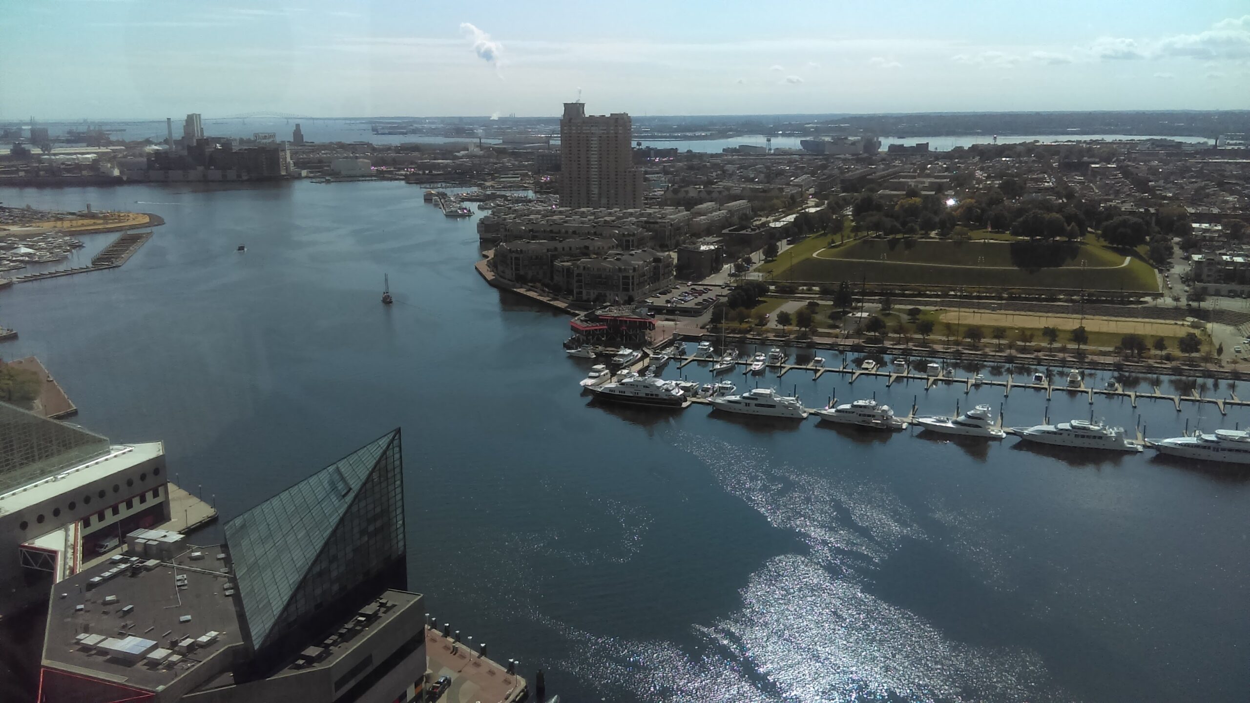 Aerial view of a harbor featuring luxury yachts at a marina, surrounded by residential buildings and green spaces, with skyline and waterway in the background.