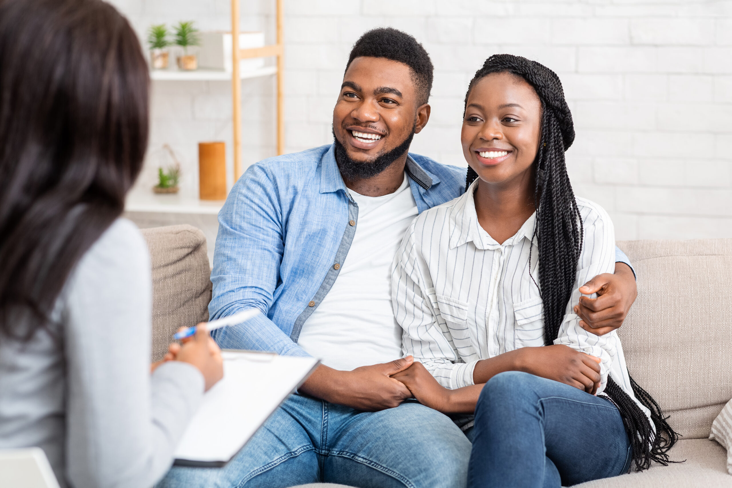 Couple smiling together during a counseling session, while a third person, presumably a counselor, takes notes. Comfort and engagement are evident.
