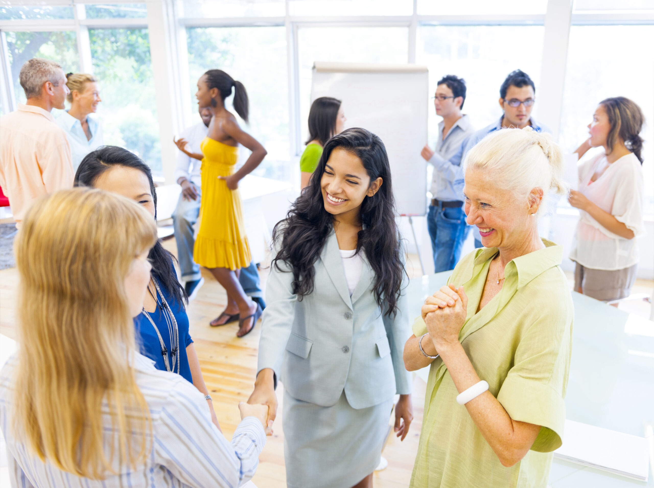 A diverse group of professionals engages in networking, with two women shaking hands in the foreground, showcasing a collaborative business environment.