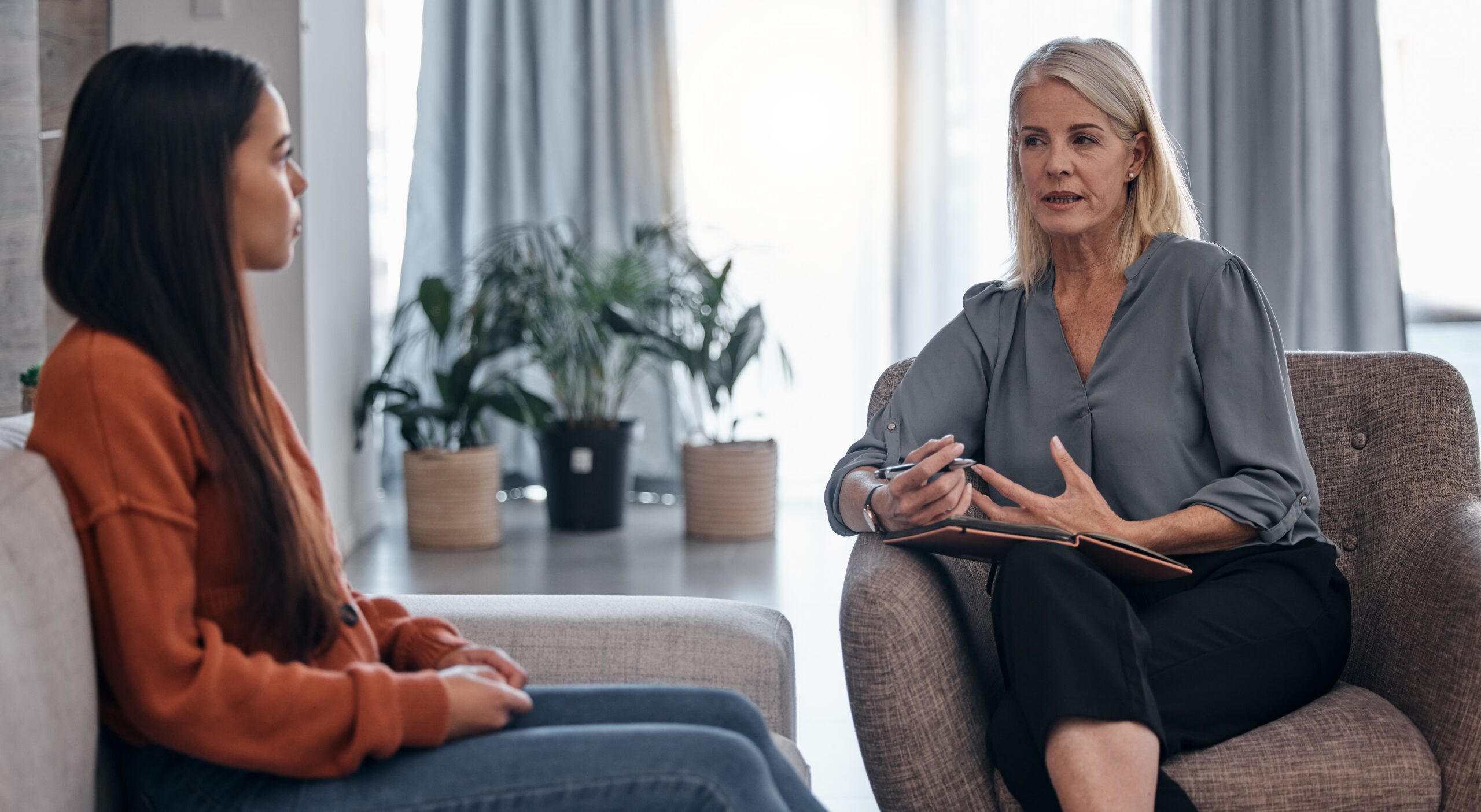 A therapist listens attentively as a young woman shares her thoughts during a counseling session in a cozy, plant-filled room.
